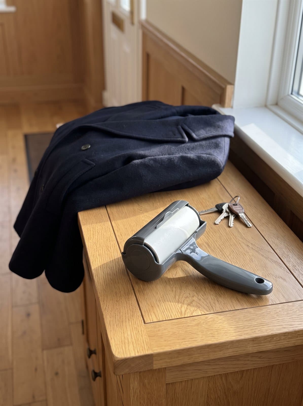 Grey lint roller on an oak hallway console beside a folded navy wool coat and a set of keys. AI-generated · Nano Banana Pro