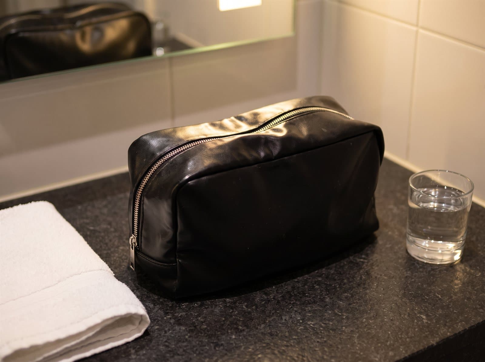 Black wash bag with a curved metal zip on a dark stone hotel bathroom counter, a folded white towel beside it. AI-generated · Nano Banana Pro